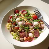 Tossed Spring Antipasto Pasta Salad with salami, pickled vegetables, and red onion, drizzled with zesty dressing on a marble surface.