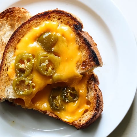 Golden, buttery sourdough grilled cheese oozing sharp cheddar and tangy pickled jalapeños on a rustic plate.  